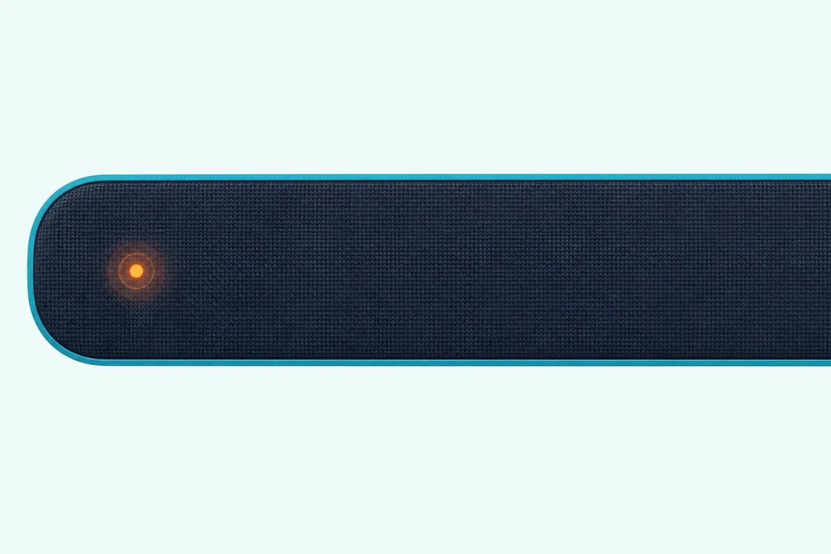 Left end of a slim soundbar in close-up, one small round light on its front glowing orange beside a plain grille